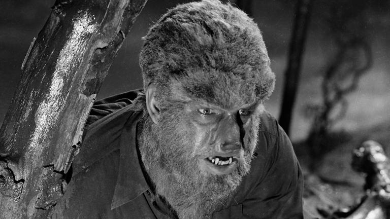 Classic black-and-white werewolf close-up with intense gaze, fur-covered face, and bared fangs beside a gnarled tree.