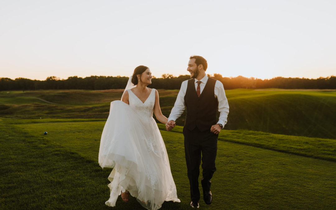 A bride and groom walking hand-in-hand across a lush golf course at golden hour, smiling warmly at each other.