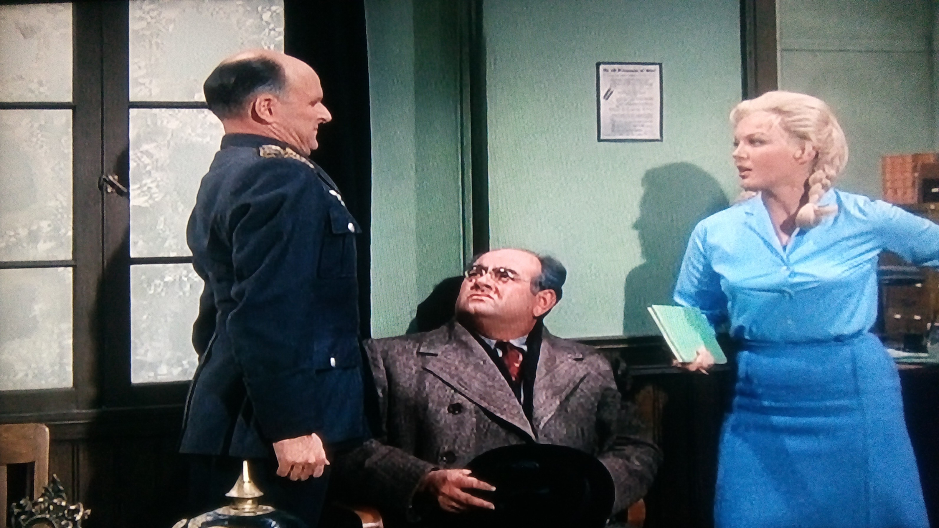 Three characters in a vintage office scene: a uniformed officer, a seated man in a coat holding a hat, and a blonde woman in blue holding a folder.