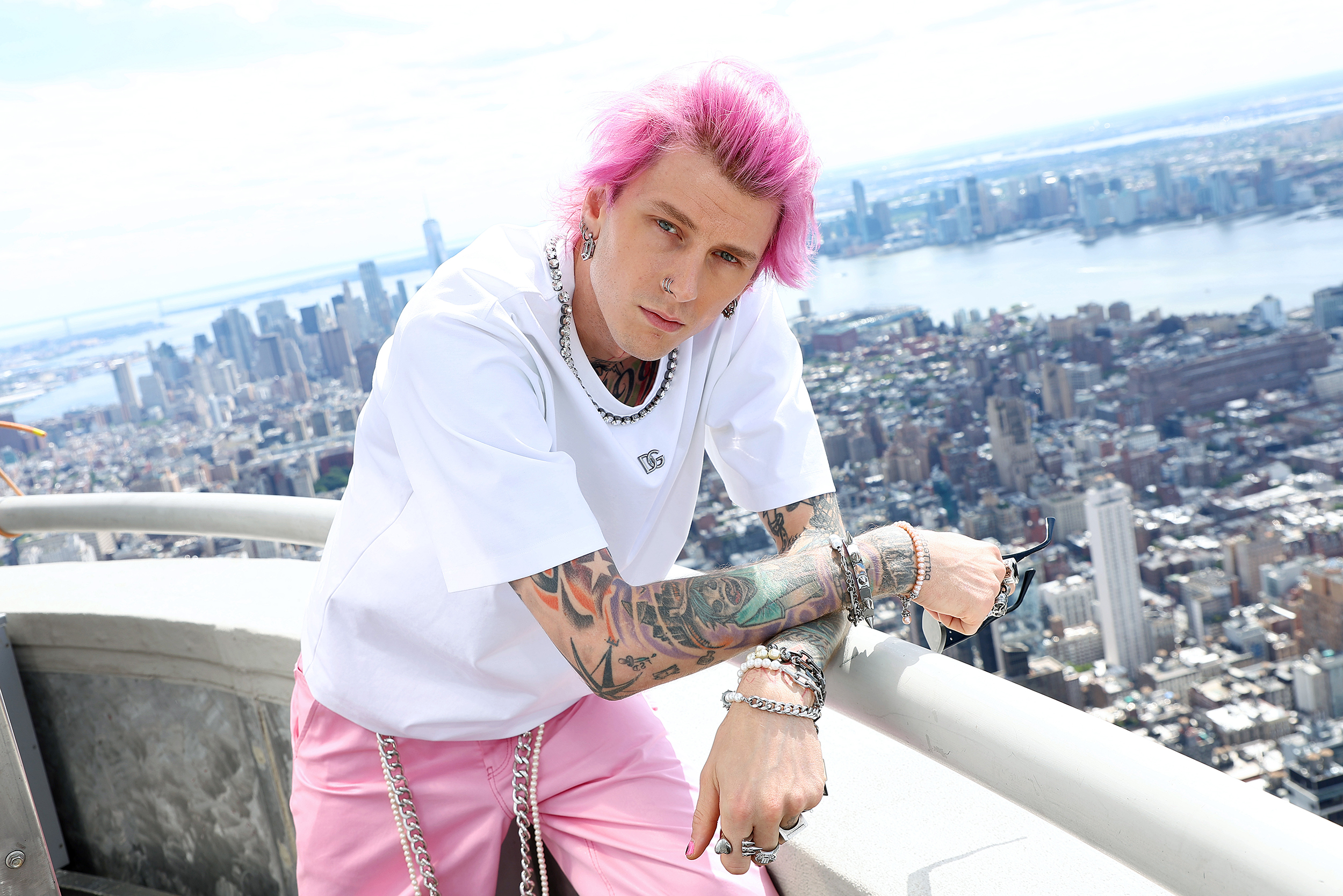 Pink-haired tattooed man in white DG shirt and pink pants poses on rooftop overlooking NYC skyline.