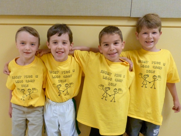 Four smiling boys in matching yellow "Saint Pius X LEGO Camp 2015" t-shirts pose together indoors.