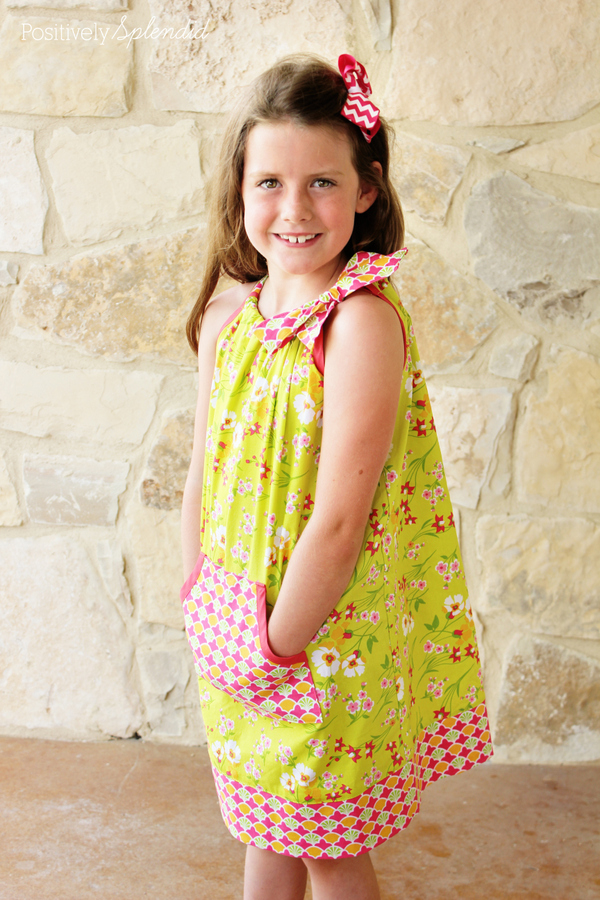 Smiling girl in a bright green floral dress with pink trim and bow, standing against a stone wall.
