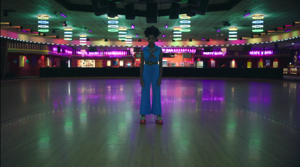 A young woman in a blue jumpsuit stands on roller skates in an empty, neon-lit roller rink at night.