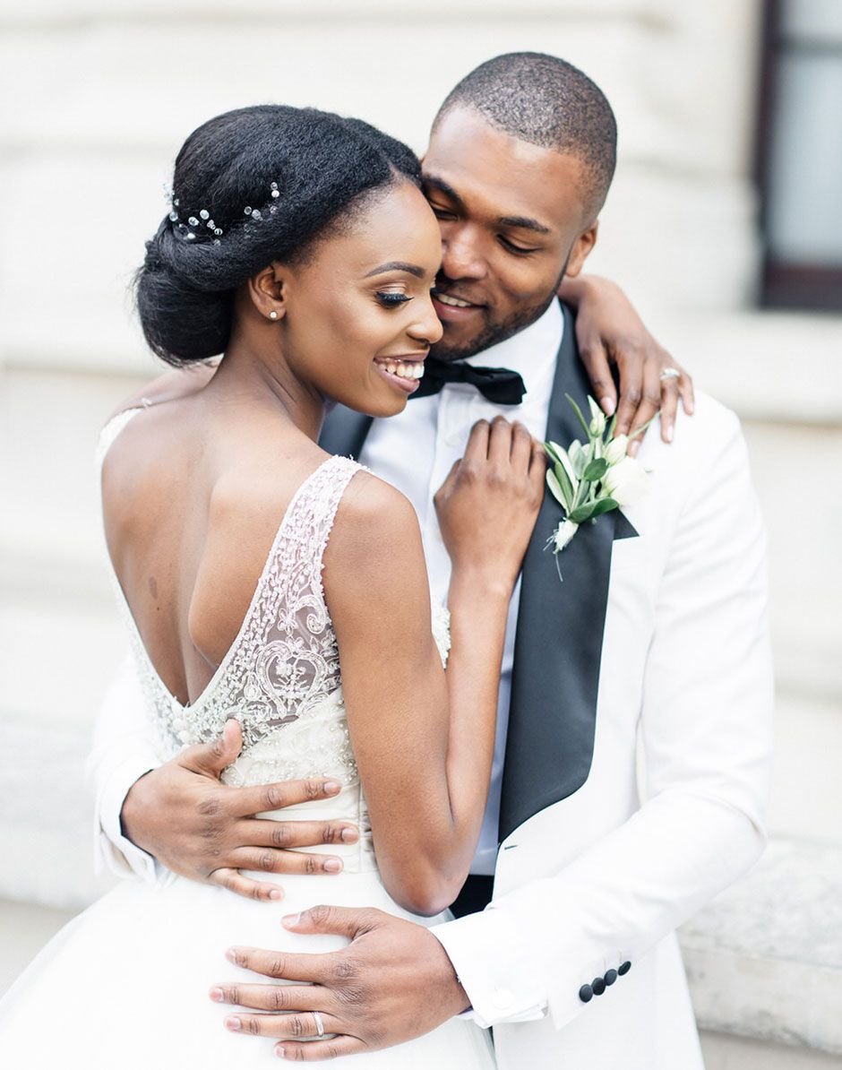 Black bride and groom in elegant white wedding attire embracing warmly outdoors.