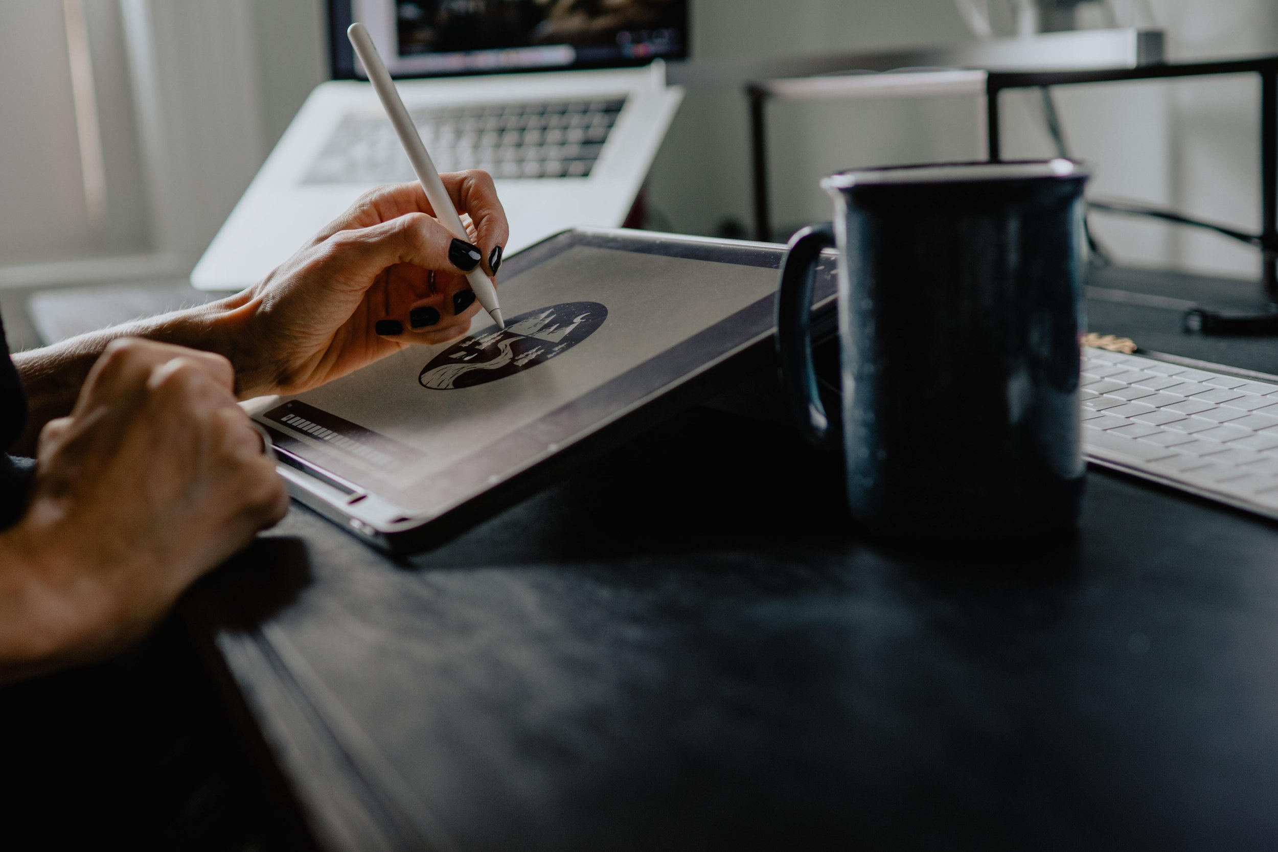 A designer uses a stylus on a tablet to draw a landscape illustration, with a laptop and coffee mug nearby on a dark desk.