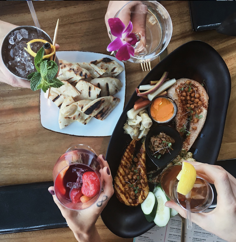 Overhead view of a vibrant mezze platter with grilled flatbread, hummus, chickpeas, and colorful cocktails on a wooden table.