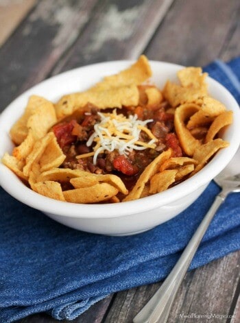 A white bowl of chili topped with shredded cheese and Fritos corn chips, set on a blue napkin over rustic wood.