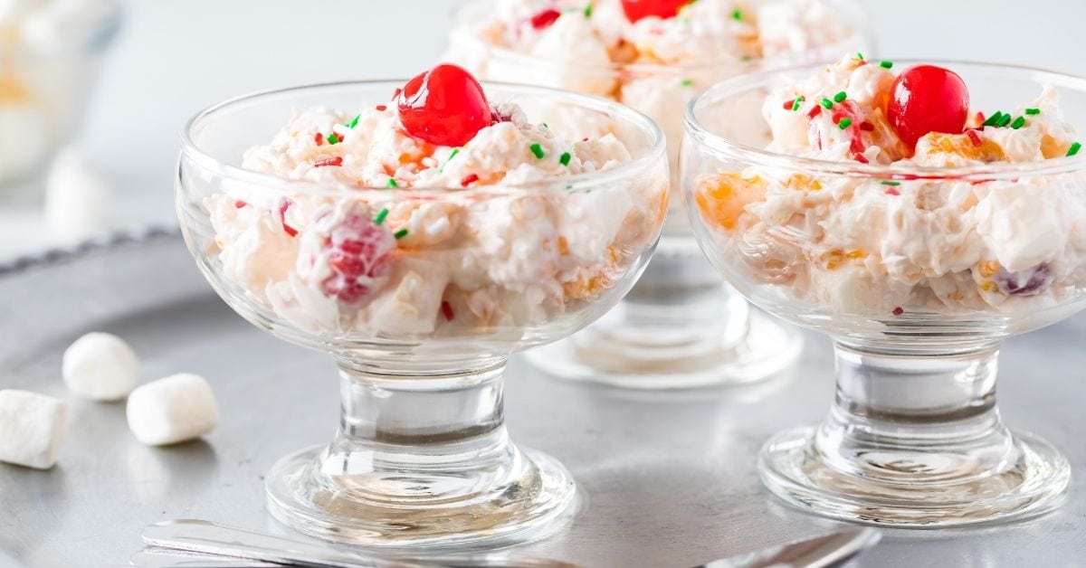 Three glass dessert cups filled with creamy ambrosia salad topped with maraschino cherries and festive sprinkles.