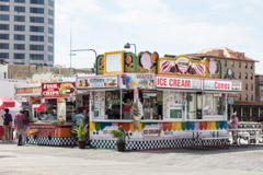 Colorful vintage-style ice cream and candy kiosk on a city plaza with pedestrians and modern buildings in background.