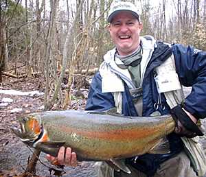 A smiling angler in winter gear holds a large, colorful rainbow trout in a wooded stream setting.