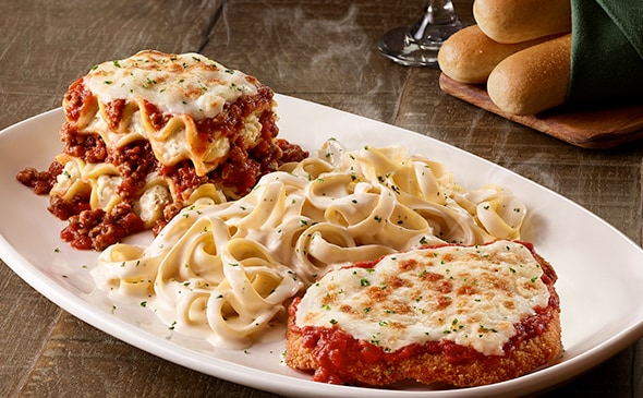 Steaming plate of lasagna, fettuccine Alfredo, and chicken Parmigiana with garlic bread on the side.