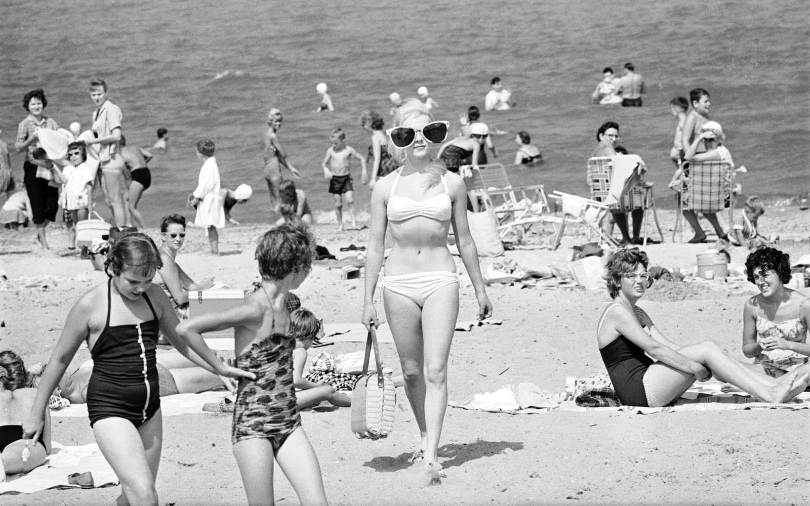 A stylish woman in a white bikini and oversized sunglasses walks confidently across a crowded 1960s beach, surrounded by families and swimmers.