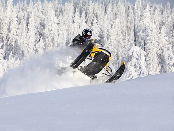 A rider in black gear maneuvers a yellow and black snowmobile through deep powder, kicking up snow against a backdrop of frost-laden evergreens.