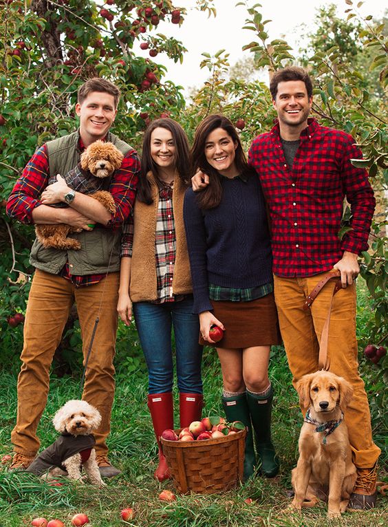 Four smiling friends with three dogs posing in an apple orchard, holding apples and a basket, dressed in autumn plaid and corduroy.