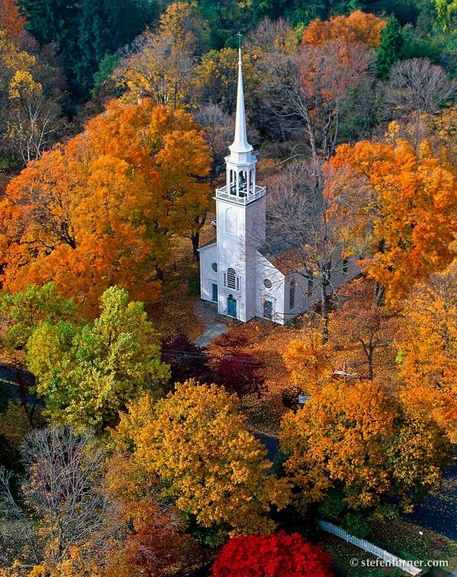 A white colonial-style church with a tall steeple nestled among vibrant autumn trees in full fall color.