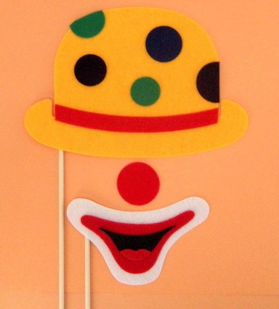 Colorful felt clown face prop with yellow polka-dot hat, red nose, and wide smiling mouth on sticks against peach background.