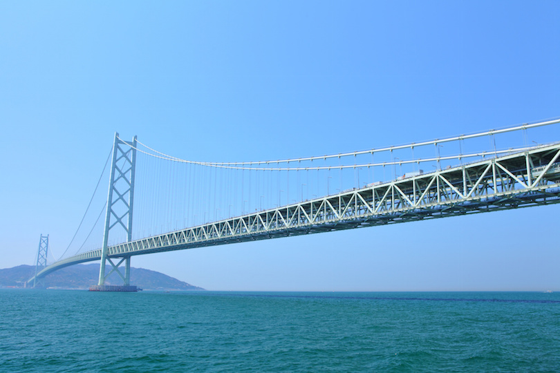 Akashi Kaikyō Bridge spanning the Seto Inland Sea under a clear blue sky, with greenish-blue waters and distant hills.