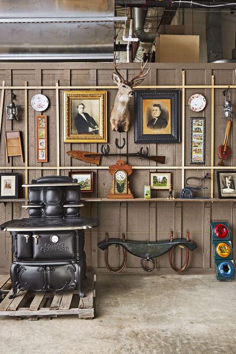 Vintage wood stove, mounted deer head, framed portraits, and antique tools displayed on rustic wooden wall shelves.