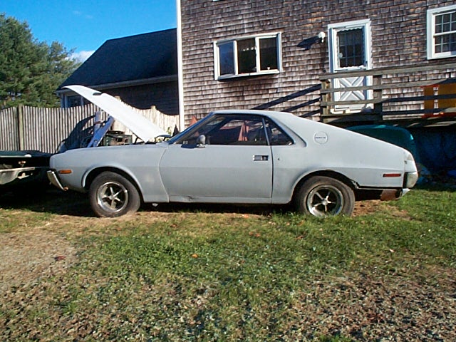Light gray 1970s AMC Gremlin coupe with hood open, parked on grass beside a wooden-shingled house.