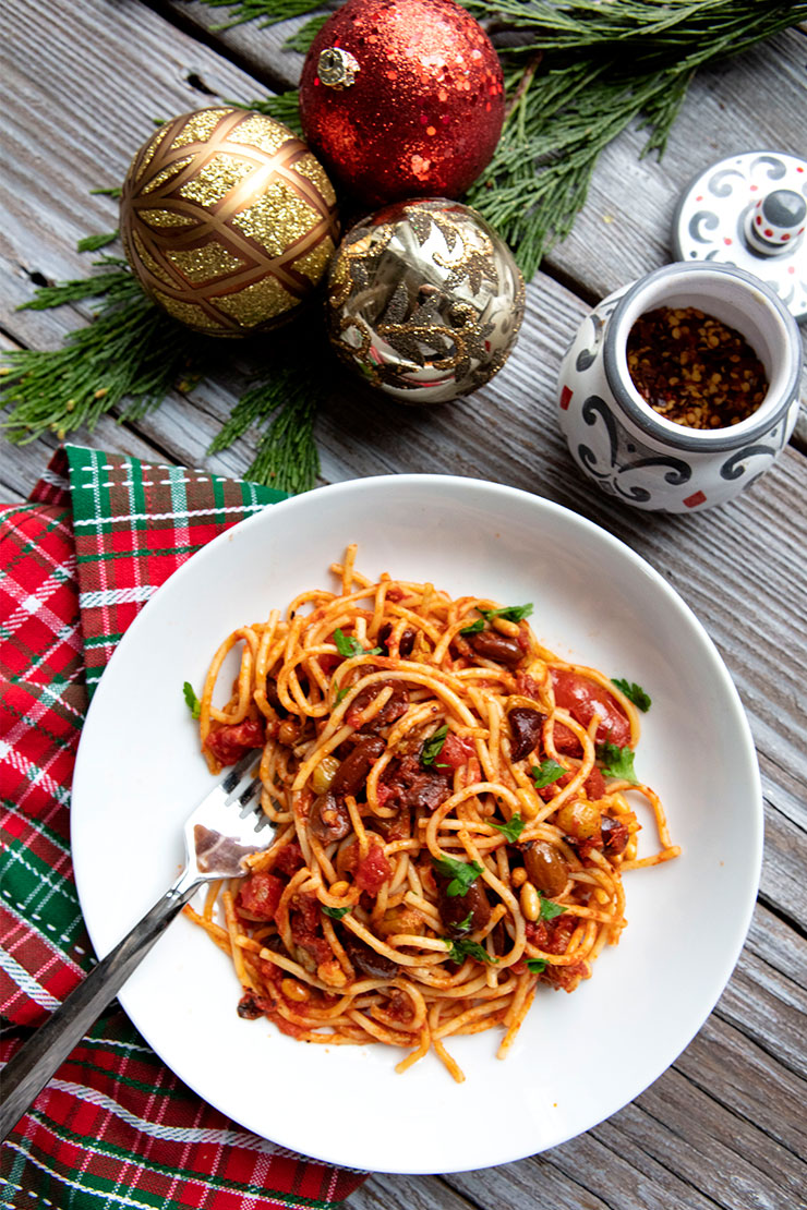 Spaghetti with tomato sauce, olives, capers, and herbs on a white plate beside Christmas ornaments and plaid napkin.