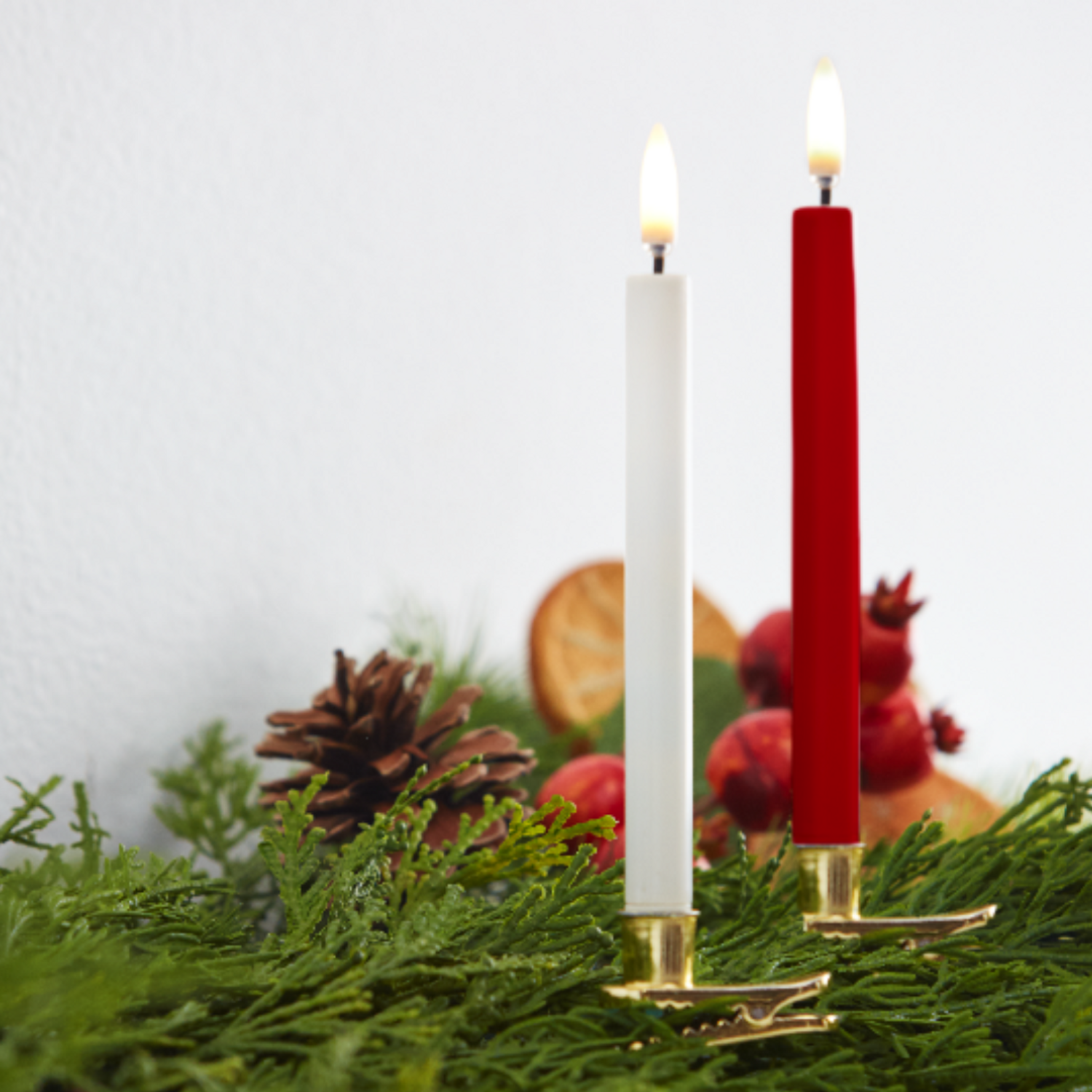 Two lit candles-white and red-in gold clip holders on evergreen garland with pinecone and dried fruit accents.