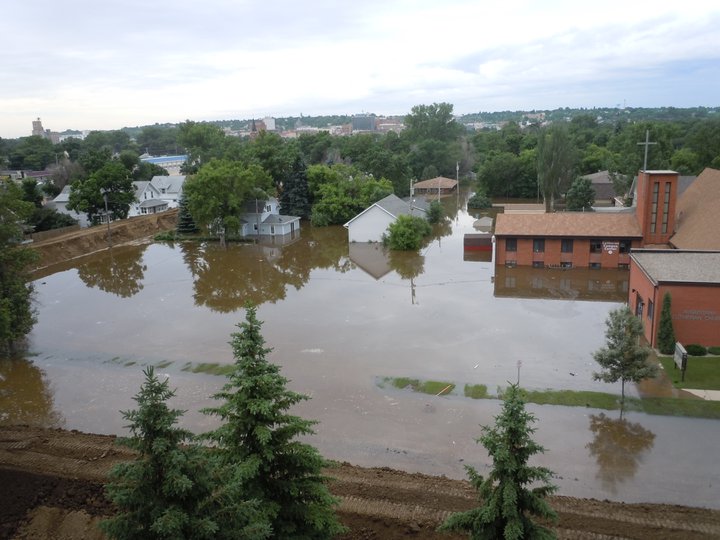 Flooded residential area with submerged houses, trees, and a church under overcast skies.