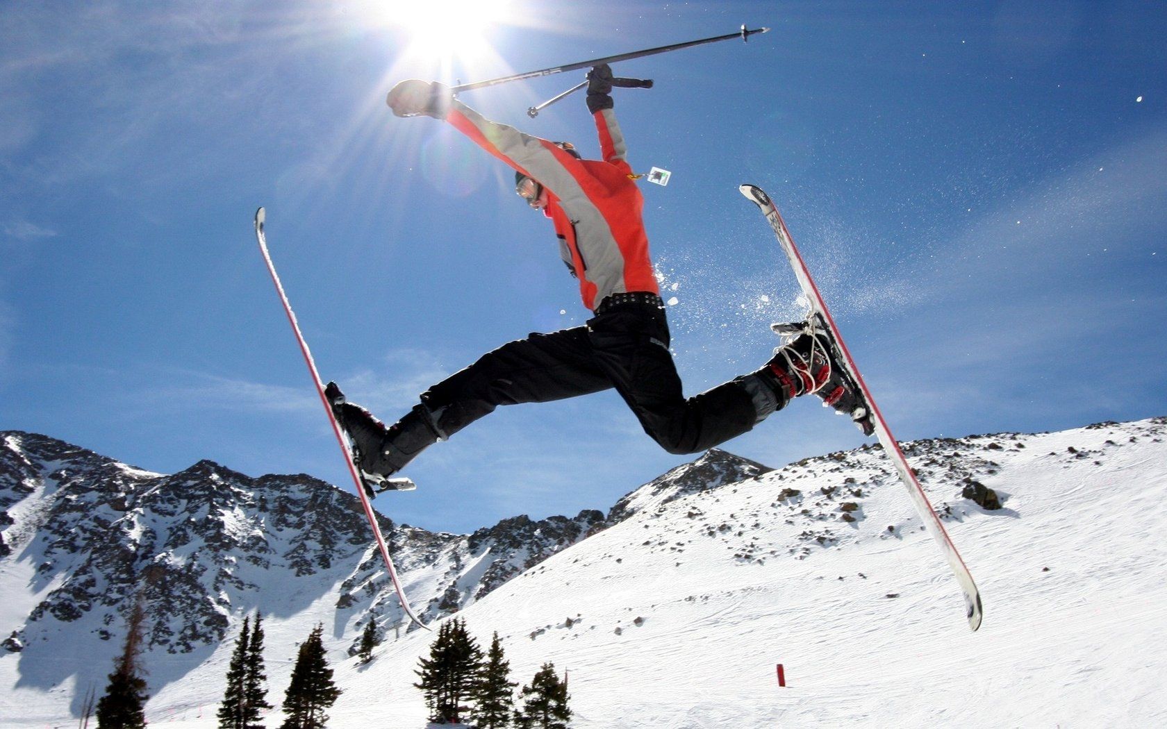 Skier in mid-air performing a jump against a sunny mountain backdrop, wearing red jacket and black pants.