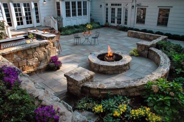 Stone patio with circular fire pit, outdoor dining set, and lush garden surrounding a white house.