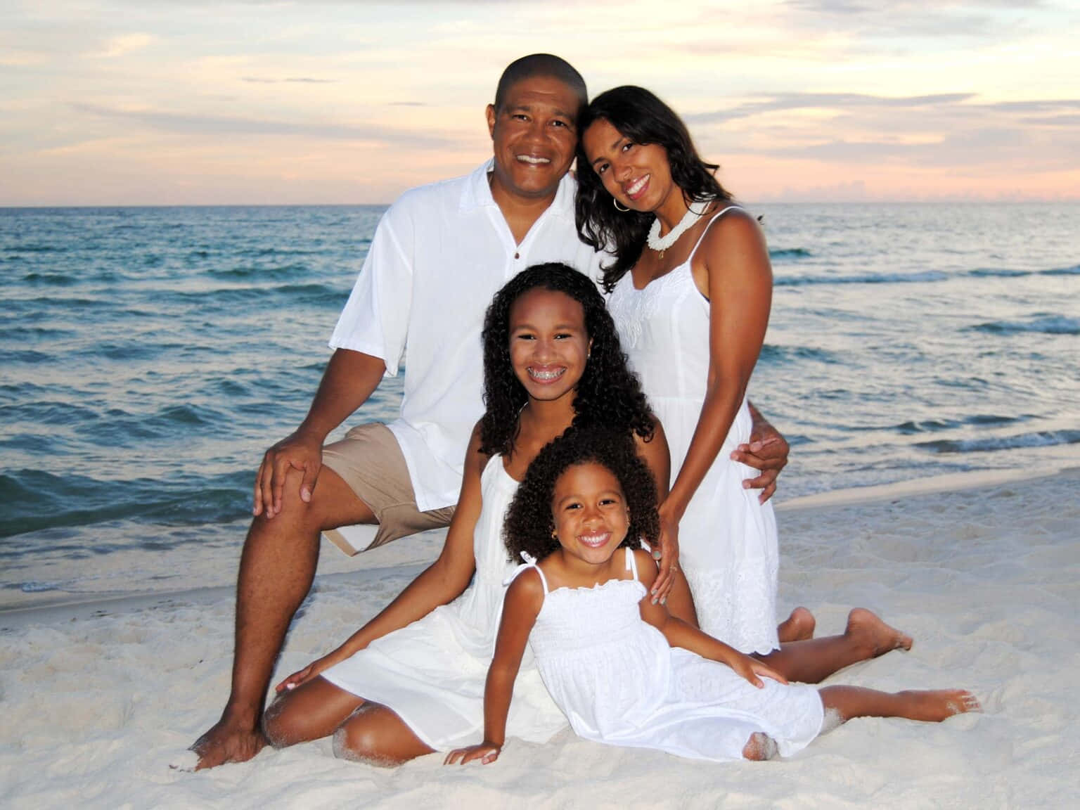 A smiling family of four in white outfits posing on a sandy beach at sunset with ocean waves behind them.