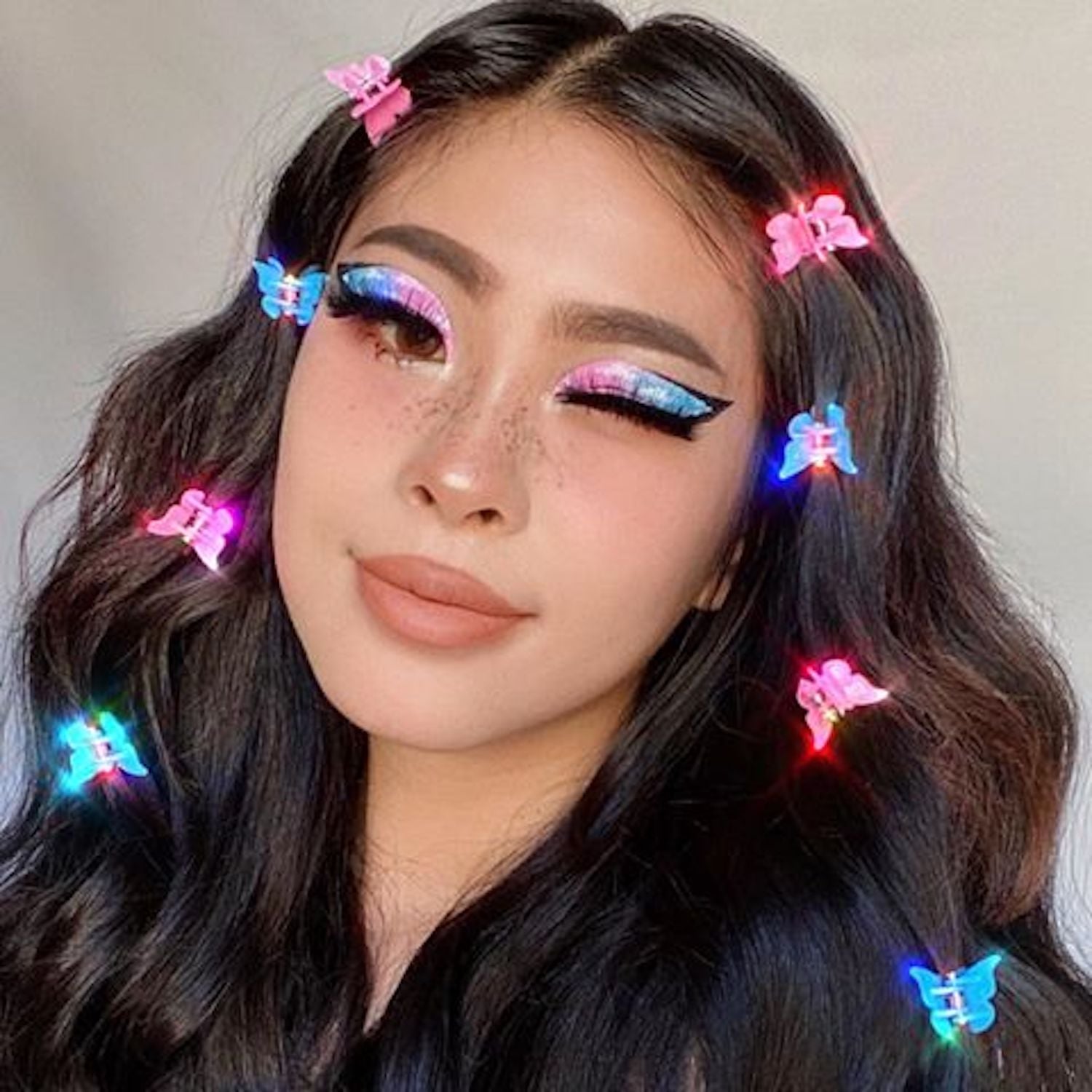 Woman with vibrant butterfly hair clips and colorful glittery eye makeup, wavy dark hair, and freckles.