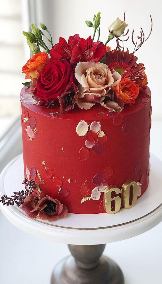 Vibrant red 60th birthday cake adorned with fresh and sugar flowers, gold leaf accents, and golden "60" numerals on a white cake stand.