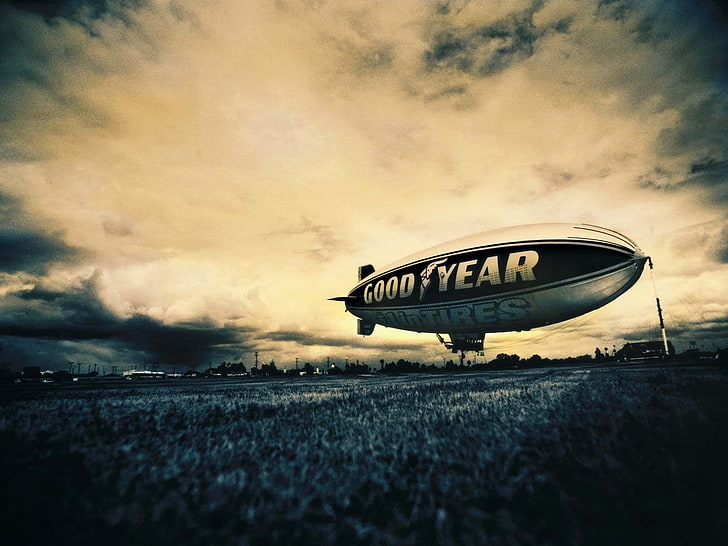 A Goodyear blimp floats low over a grassy field under a dramatic, cloudy sky at dusk.