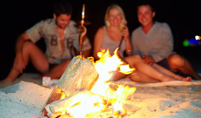 Three friends smiling around a crackling campfire on a sandy beach at night.