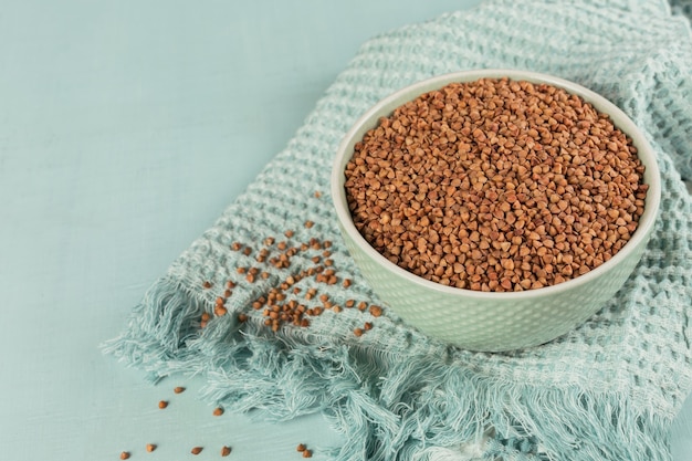 A light green ceramic bowl filled with raw buckwheat groats, resting on a textured teal cloth with scattered grains nearby.