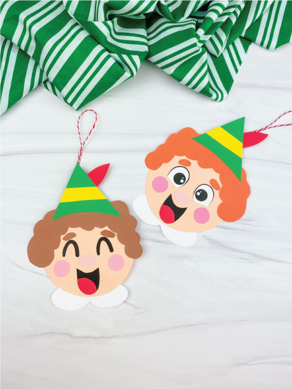 Two cheerful paper elf ornaments with green-yellow hats, rosy cheeks, and red-and-white string hangers on a white marble surface.