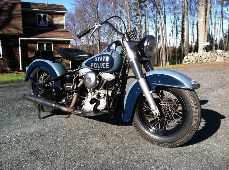 Vintage blue-and-silver 1940s Harley-Davidson police motorcycle parked on gravel driveway near a house and woods.