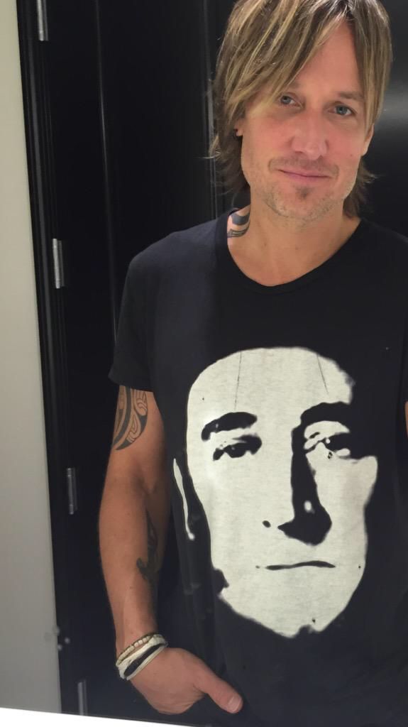 Keith Urban wearing a black T-shirt with a stylized white face print, showing tattoos and bracelets, posing indoors.