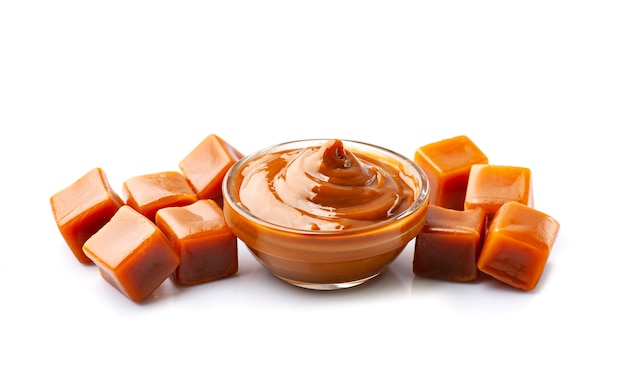 Rich caramel sauce in a glass bowl surrounded by glossy caramel cubes on white background.