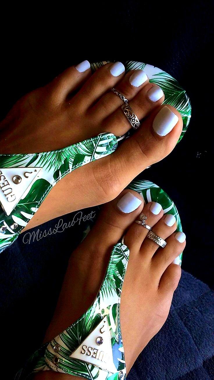 Feet in white-polished nails wearing green palm-print Guess flip-flops with silver toe rings, against dark background.
