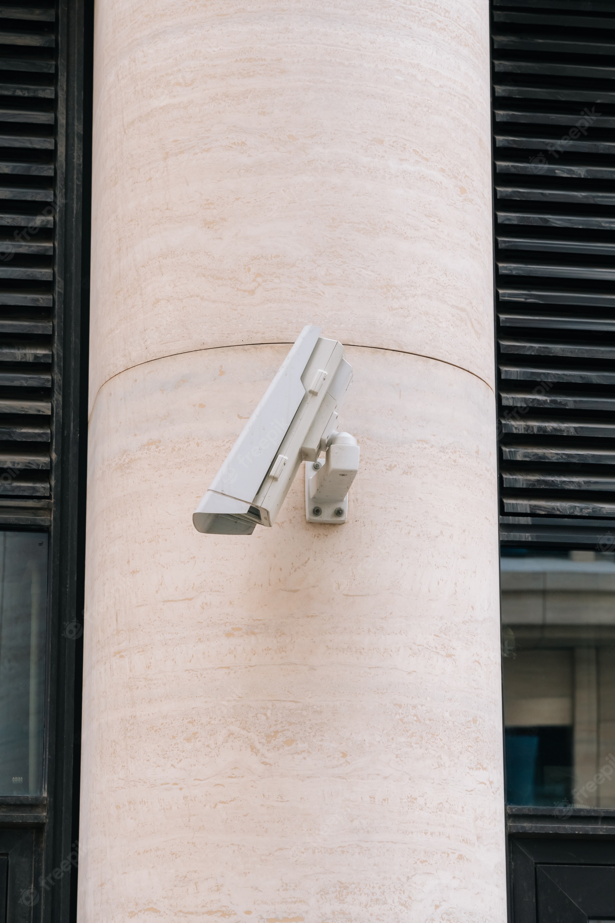 White security camera mounted on a beige stone column beside black slatted panels.