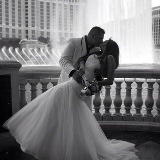 A black-and-white wedding photo of a couple embracing passionately in front of a fountain with a city skyline behind them.