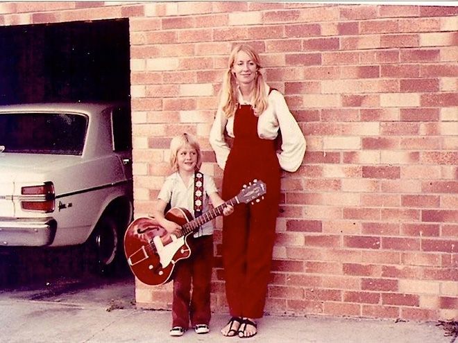 A woman and young child pose with a red electric guitar beside a brick wall and vintage car, 1970s style.