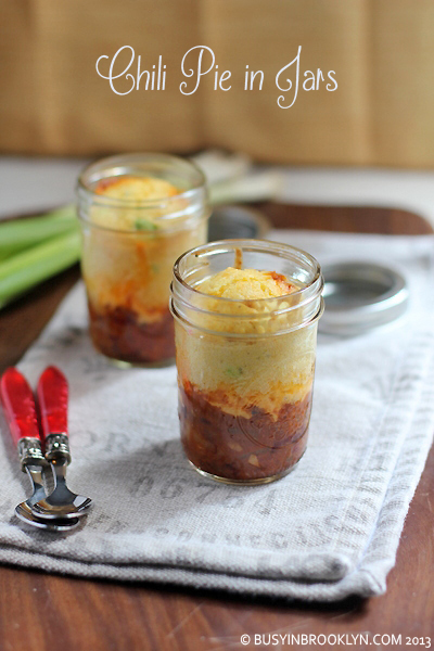 Two glass mason jars filled with layered chili pie-chili base, cheese, and cornbread topping-on a linen napkin with red-handled spoons nearby.