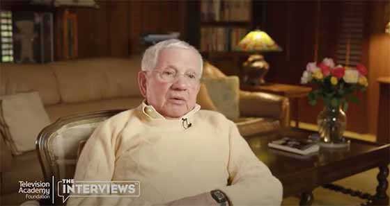 Elderly man in cream sweater seated in a warmly lit library-style room, speaking during an interview.