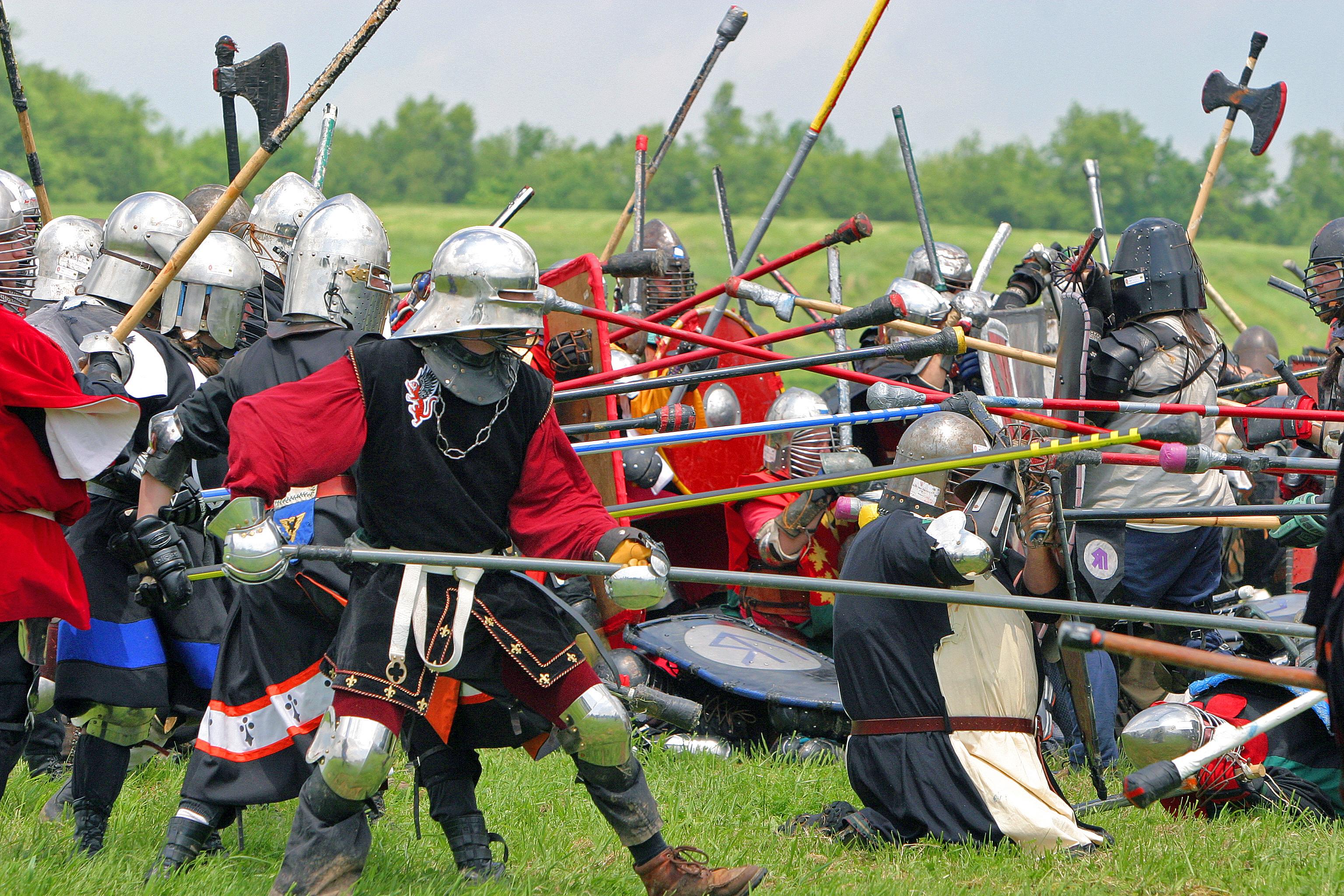 Medieval reenactors in armor clash with polearms and shields during a live combat demonstration on grassy field.