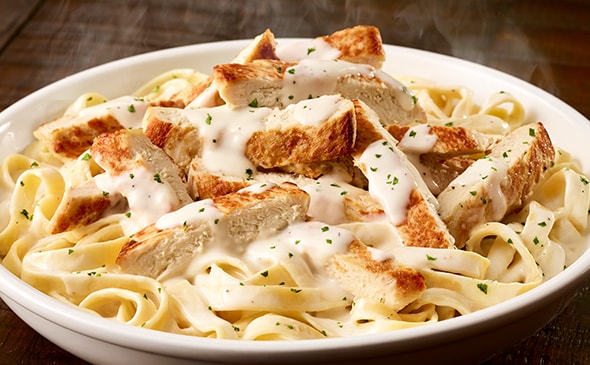 Steaming fettuccine Alfredo topped with grilled chicken pieces and fresh parsley in a white bowl.