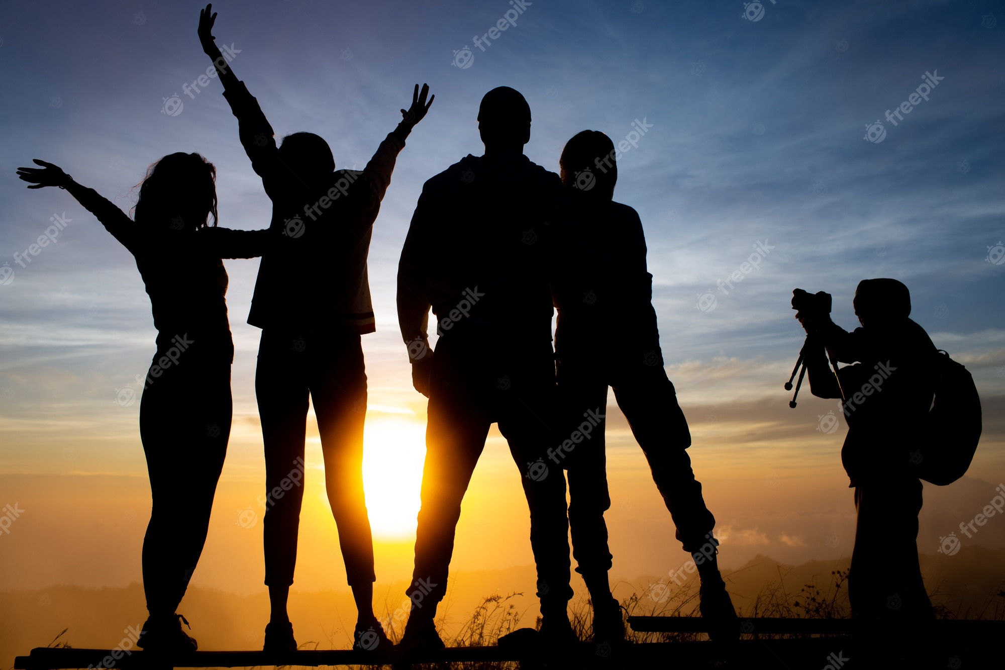 Silhouettes of five friends celebrating at sunrise on a mountain ridge, one photographing the moment.