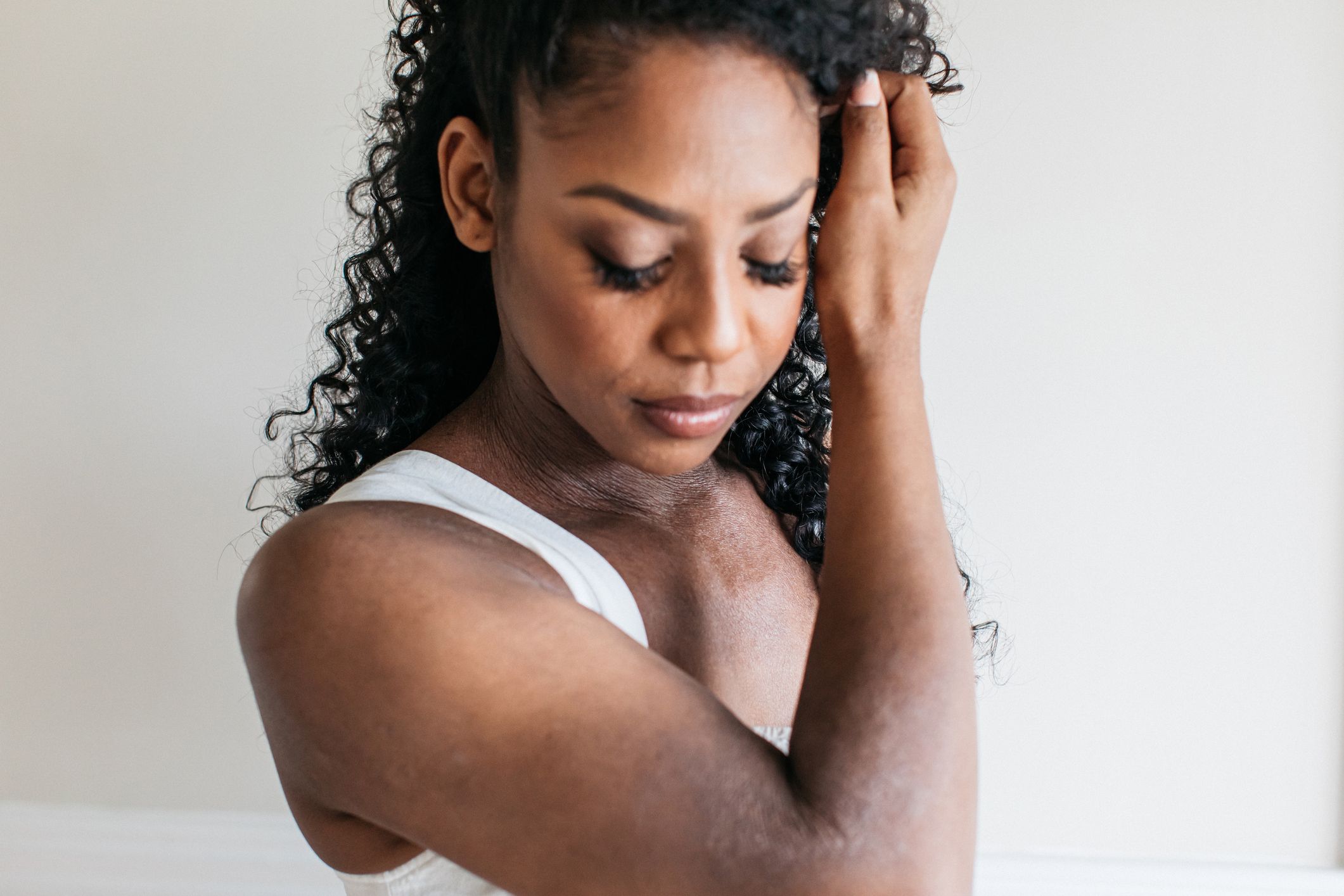 Black woman with curly hair gently touching her temple, wearing white tank top, soft natural lighting.