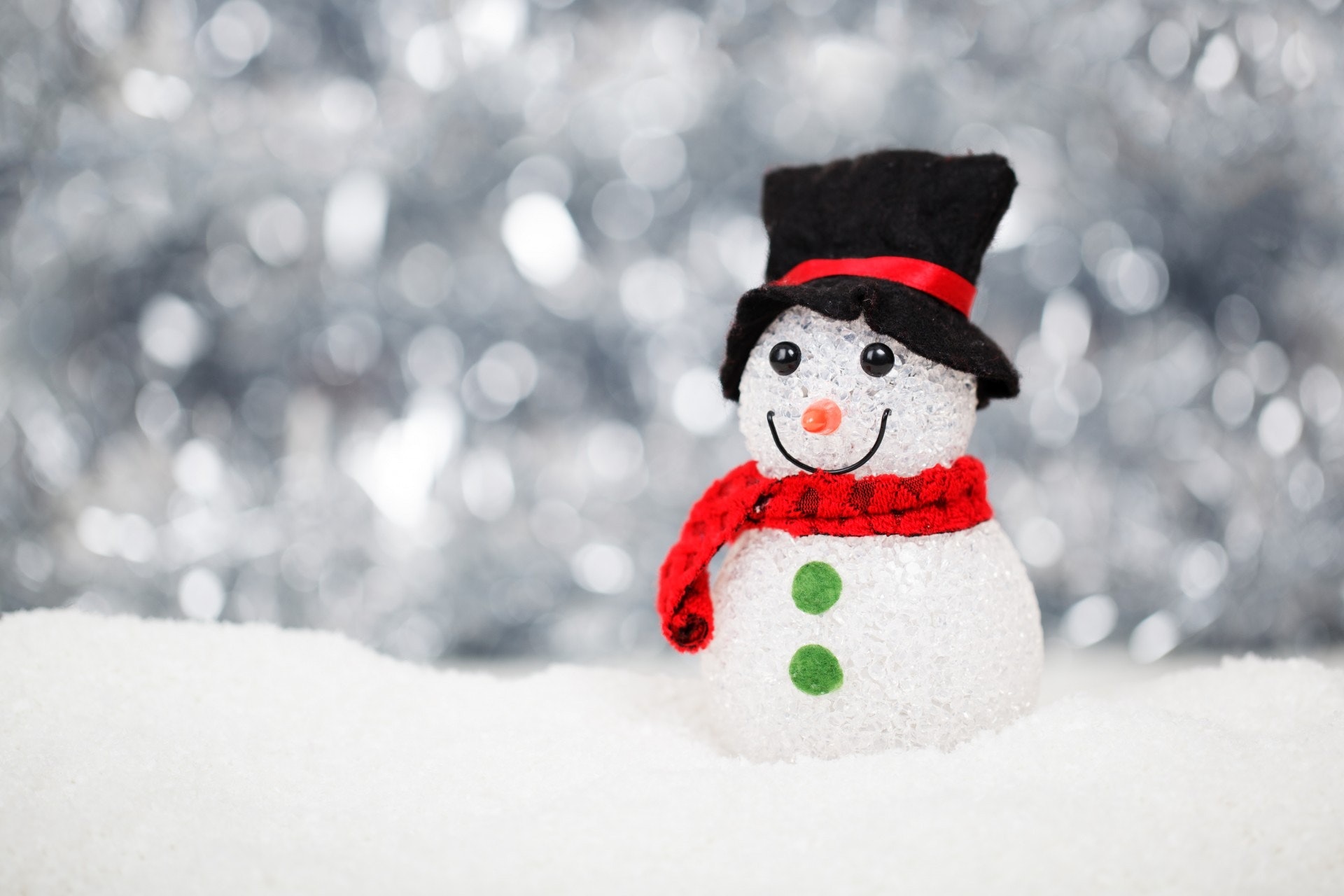 Sparkling white snowman with black top hat, red scarf, green buttons, and orange nose on snowy surface against silver bokeh background.