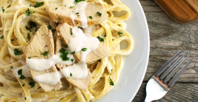 Creamy fettuccine Alfredo with sliced grilled chicken, parsley garnish, served on a white plate beside a fork on rustic wood.